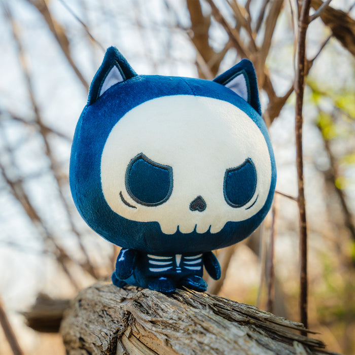 Cloudheads: 5" Skeleton Cat (Skelly) is a cat skeleton Cloudheads plushie. The skull face has a grumpy look to it. It's sitting on a log and there are bare trees in the background.