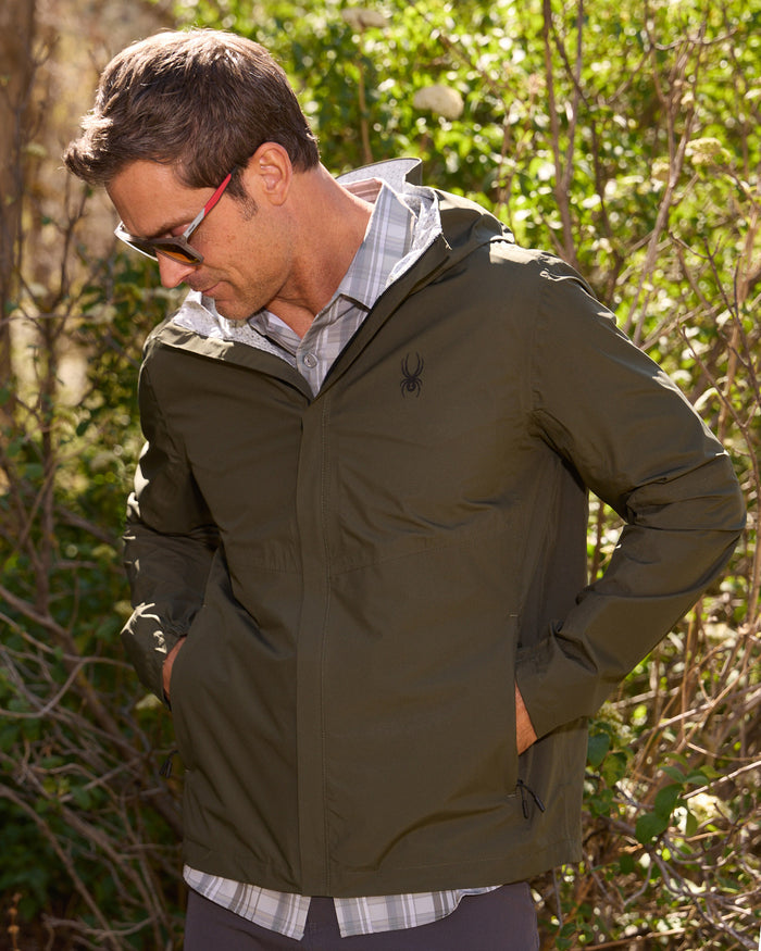 Man wearing a green jacket outdoors with greenery in the background