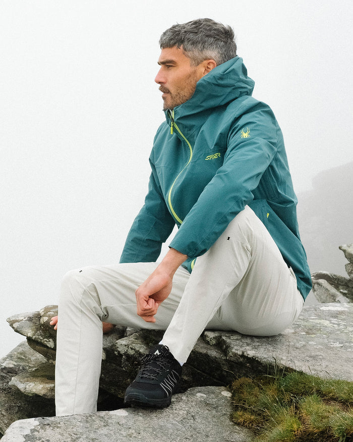 Man wearing a green jacket sitting on rocks with a mountainous background