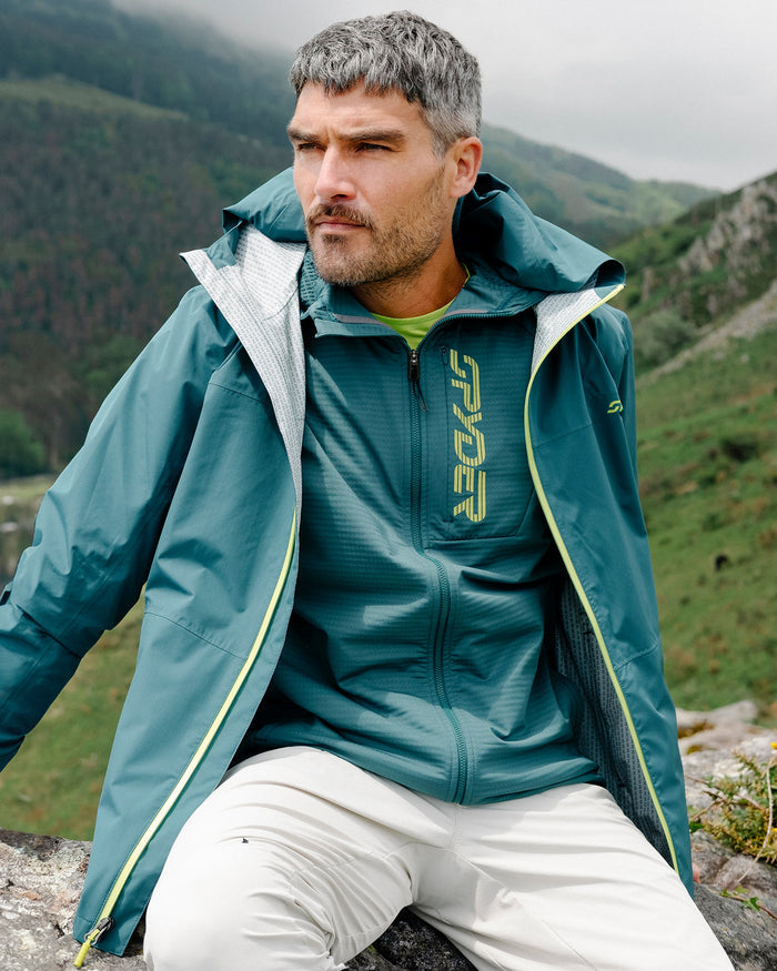 Man wearing a green jacket with a brand logo, sitting outdoors in a mountainous area.