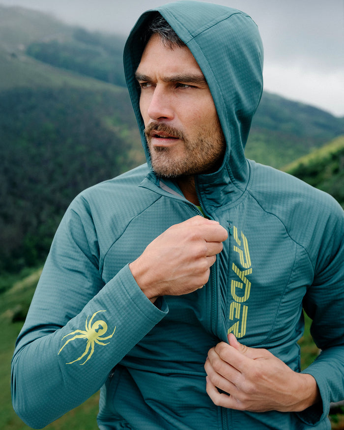 Man wearing a teal jacket with a logo in a mountainous landscape