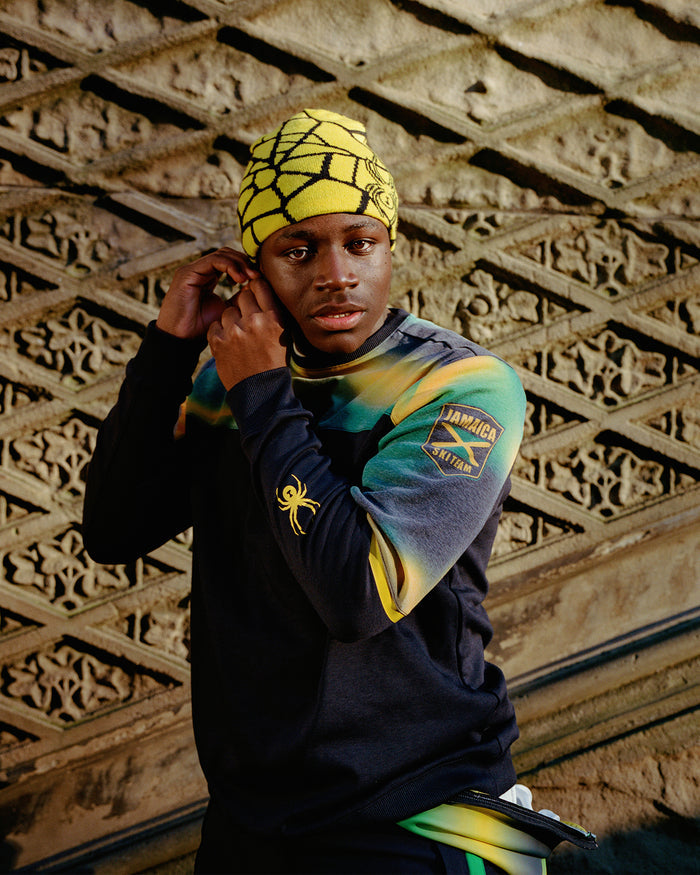 Person wearing a colorful outfit with a patterned hat against an ornate wall.
