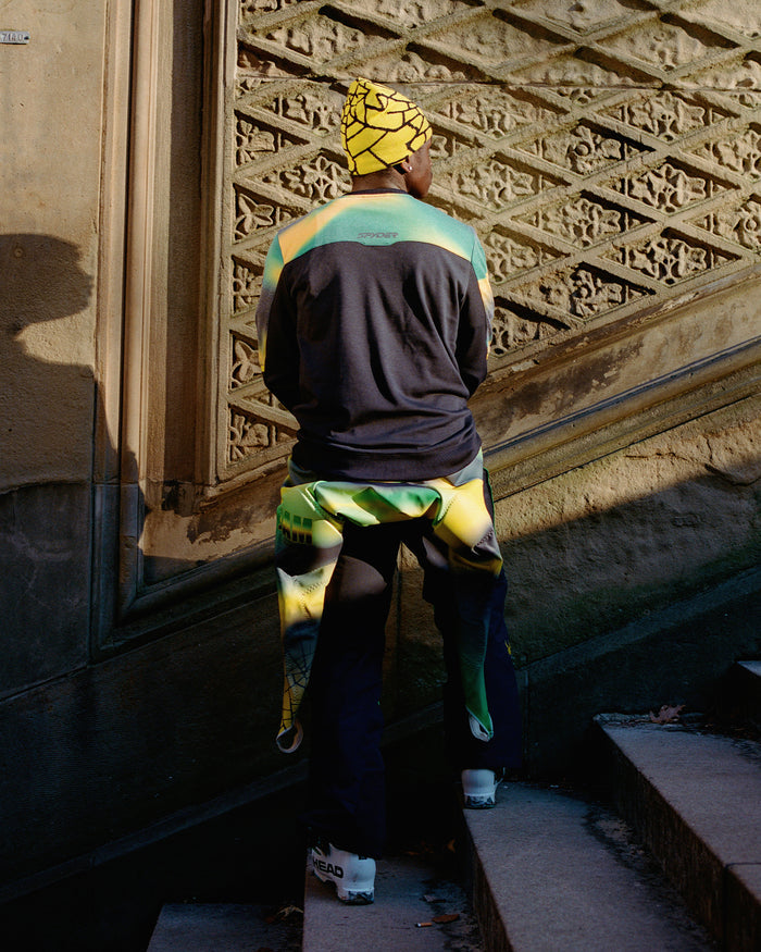 Person wearing a colorful outfit with a patterned hat and scarf, standing in front of an ornate wall.