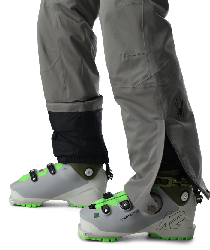 Gray ski boots with green accents worn by a person in gray pants on a white background