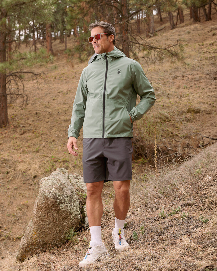 Man wearing a green jacket and gray shorts standing in a forested area.