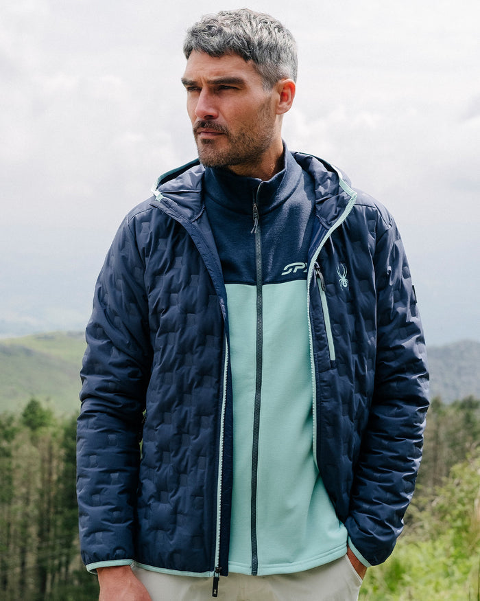 Man wearing a blue quilted jacket with a brand logo, standing outdoors with a forest in the background.