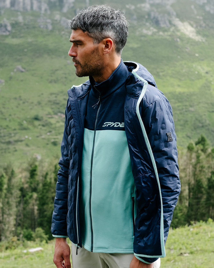 Man wearing a blue jacket with a brand logo in a mountainous landscape