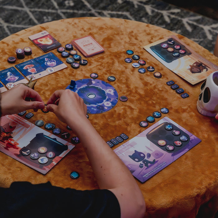 Mystic Moon Alchemy Expansion - the components of the game including resource tokens, cards and player boards are laid on a table covered in a tan velvet cloth. You can see a person's hands holding one of the tokens.